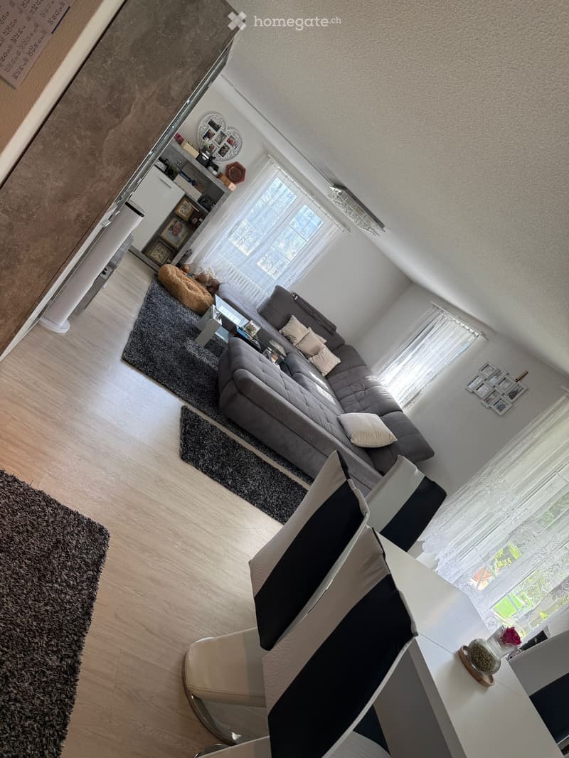 A modern living room with a grey sectional sofa, coffee table, white chair, and a small table with a cup on it. The room features large windows with white curtains and a wooden floor.