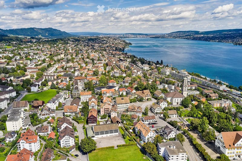 Aerial view of a town with numerous houses, buildings, and a lake in the background. The town is surrounded by green spaces and mountains.