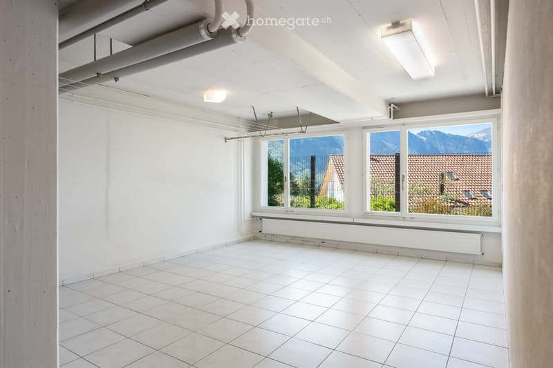 Empty room with white tiled floor, white walls, large windows showing a view of mountains and a neighboring building, and exposed ceiling pipes.
