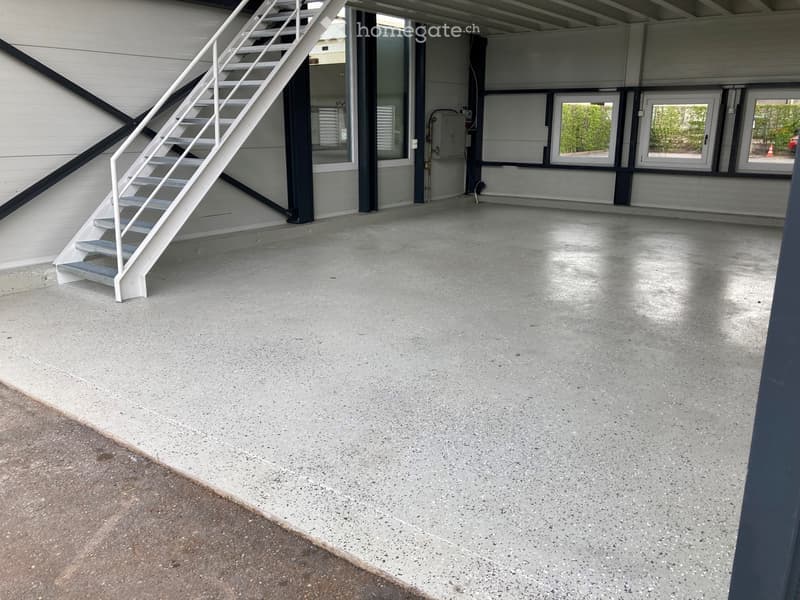 Empty garage with concrete floor, white metal staircase leading up, large windows, and industrial-style doors.