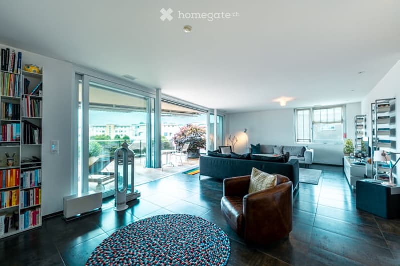Modern living room with large windows offering a view, featuring a grey sofa, leather armchair, coffee table, bookshelf, floor lamp, and a colorful rug on the dark wooden floor.