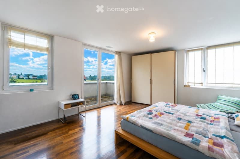 Bright bedroom with wooden flooring, large windows, balcony access, bed with colorful quilt, nightstand, and wardrobe.
