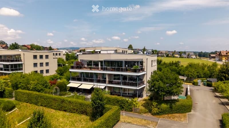 A modern multi-story residential building with balconies and large windows, surrounded by greenery and other structures in the background.