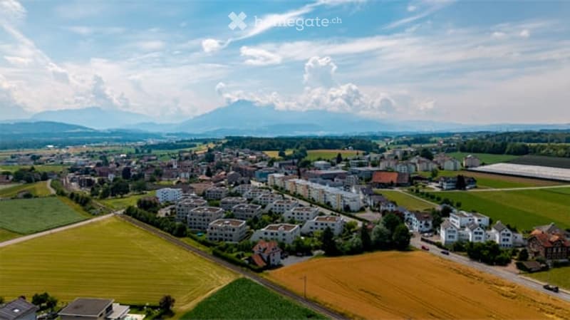 Aerial view of a suburban town with houses, green fields, and distant mountains under a partly cloudy sky.
