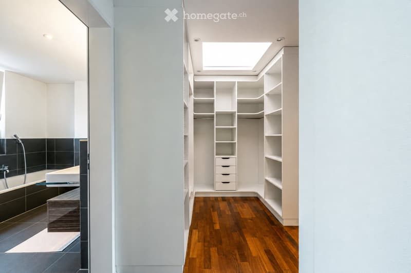 Modern bathroom with a bathtub, sink, and built-in wardrobe with shelves and drawers. The room features a wooden floor and a skylight above the wardrobe.