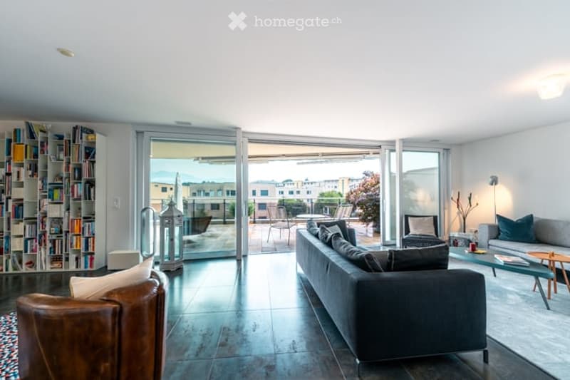 Modern living room with large glass sliding doors leading to a balcony, grey sofa, coffee table, armchair, bookshelf, and table lamp.