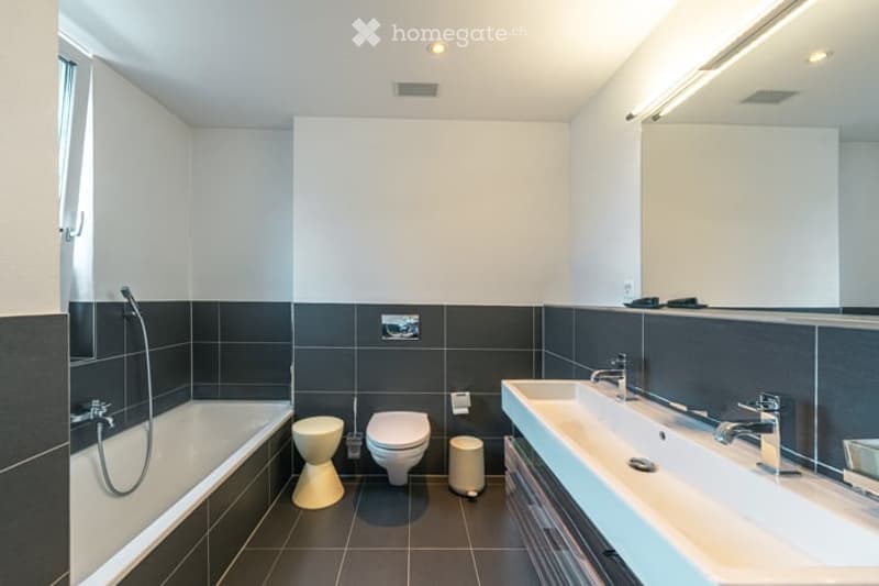 Modern bathroom with black tiled walls and floor, white fixtures including a bathtub, toilet, and double sink with chrome faucets.
