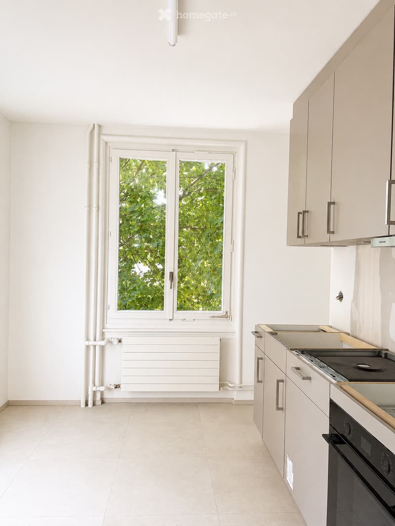 Modern, minimalist kitchen with white cabinets, a built-in oven, and a stove. Features a large window with a view of greenery, a radiator below the window, and a tiled floor.