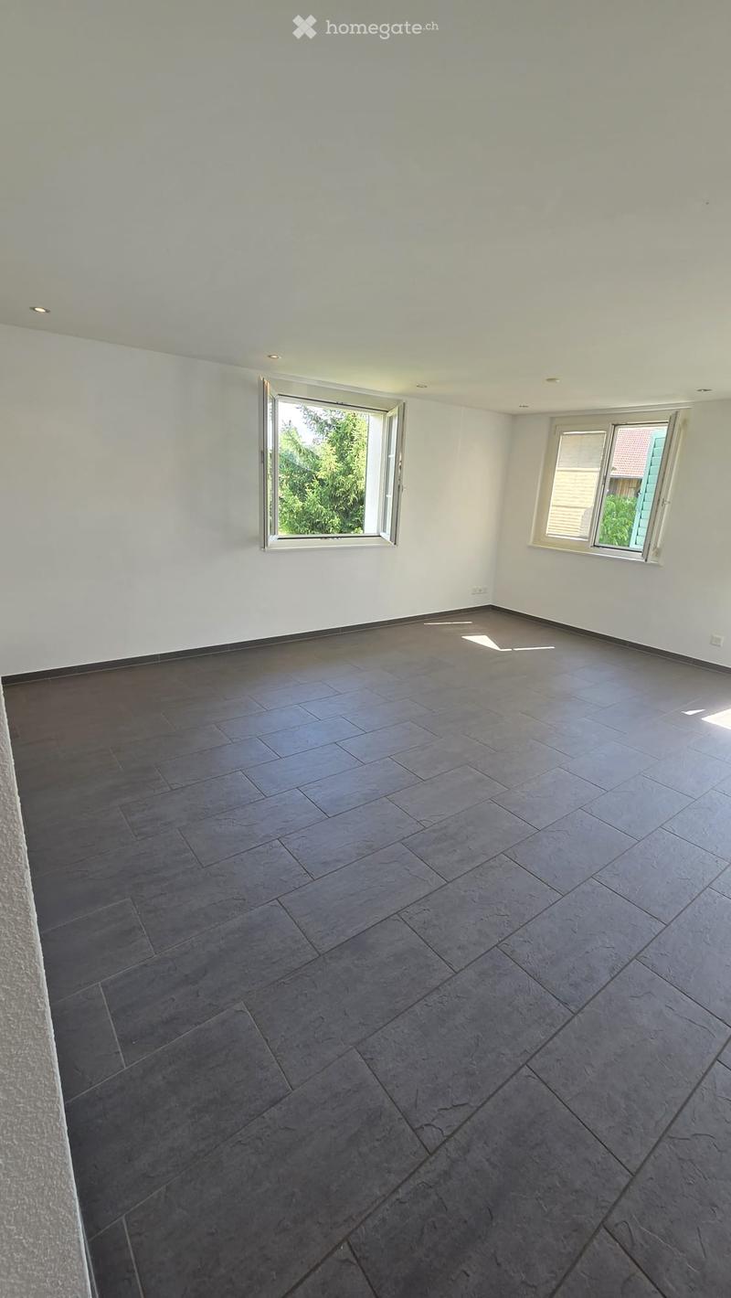 Empty room with grey tiled floor, white walls, and two windows, one of which is open to show greenery outside.