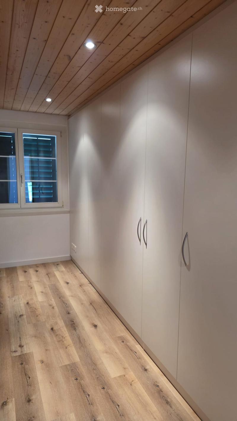 A narrow hallway with light wooden flooring, white walls, built-in white cabinets with silver handles, and a small window with blinds on the left side.