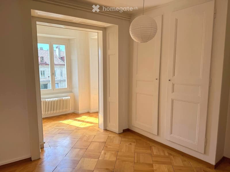 Empty hallway with wooden flooring, white walls, and a hanging spherical light fixture. A doorway leads to a room with large windows and a radiator.
