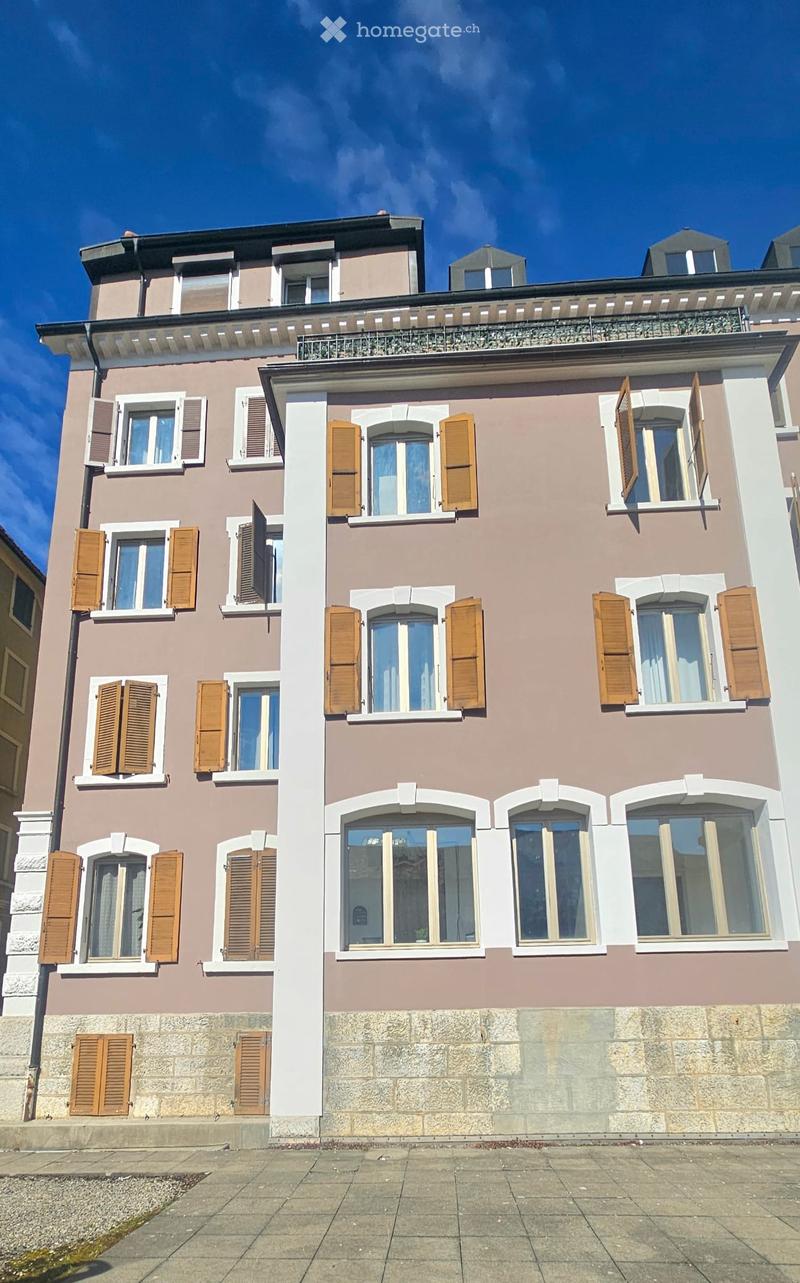 A three-story residential building with beige and brown facade, multiple windows with wooden shutters, and a balcony on the top floor.