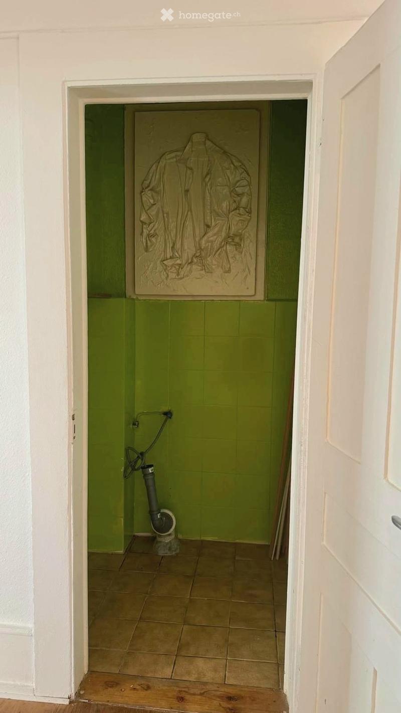 Empty room with green tiled walls, beige tiled floor, and a partially visible white door frame. A small pipe and a piece of artwork are visible.