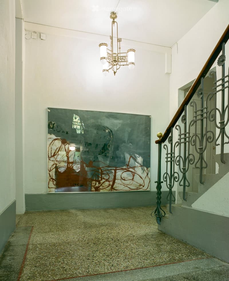Hallway with a staircase, ornate metal railing, chandelier, and a large abstract painting on the wall.