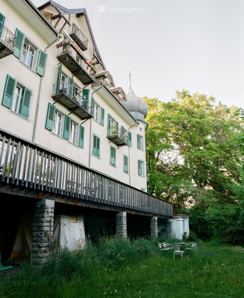A multi-story residential building with white walls, green shutters, and multiple balconies. The structure features a distinctive dome-shaped roof and is surrounded by lush greenery and a small seating area with chairs and a table.