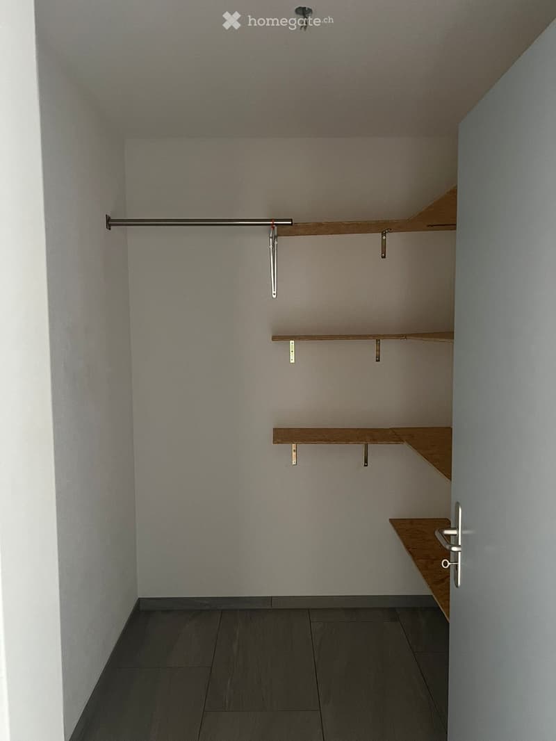Empty closet with wooden shelves and a metal rod for hanging clothes. The room has a tiled floor and white walls.