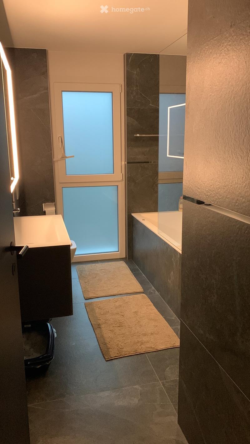 Modern bathroom with white walls and grey flooring, large mirror, double vanity, walk-in shower, and two rugs