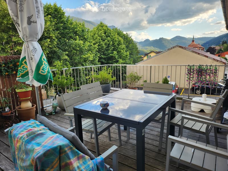 Balcony with wooden decking, glass table, chairs, umbrella, plants, and a small toy turtle.