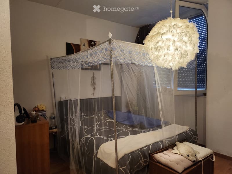 Cozy bedroom with a large bed under a decorative canopy, nightstand with lamp, and a small stool with a decorative pillow.