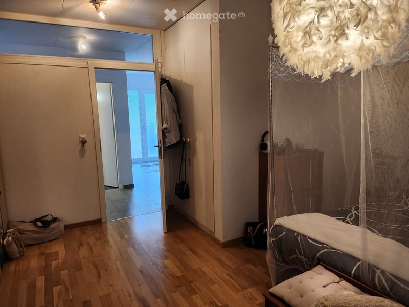 A modern bedroom with a wooden floor, a large bed with a canopy, and a nightstand. A coat and handbag are placed near the door, which leads to another room.