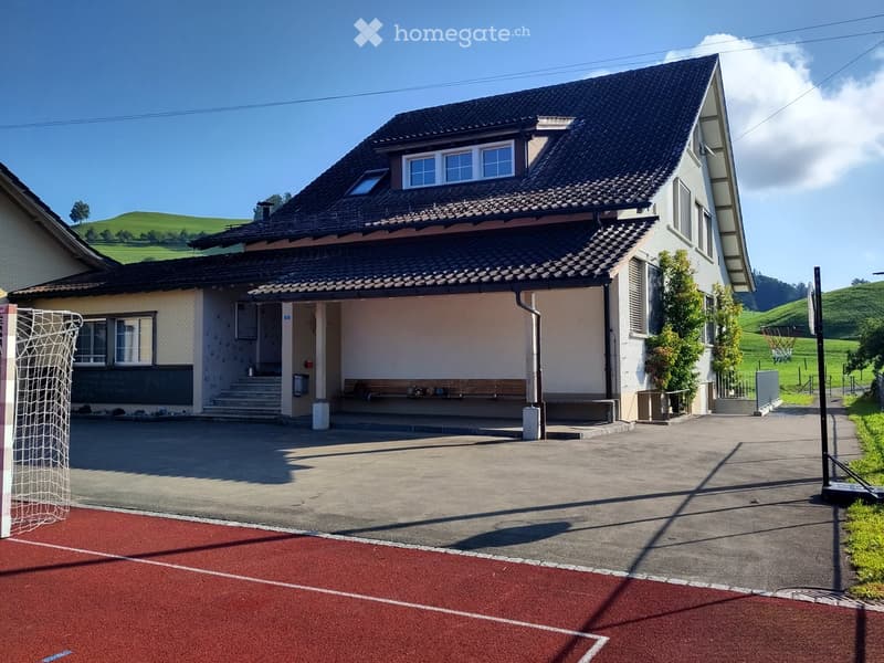 Two-story house with a dark tiled roof, multiple windows, and a covered entrance area. The house is surrounded by a paved driveway and a small sports field with a goalpost.