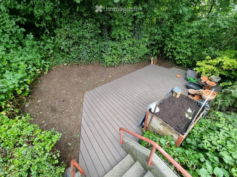 Wooden deck with a small raised garden bed, surrounded by greenery and plants. Includes gardening tools and pots.