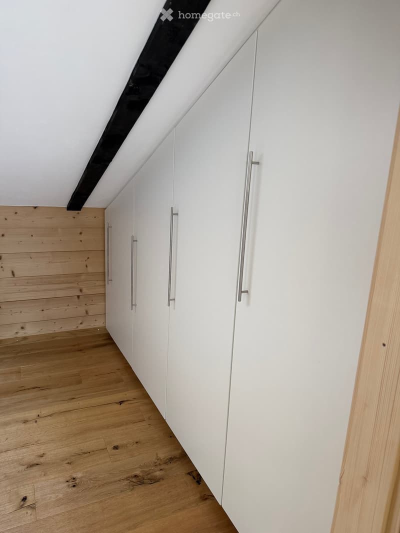 An empty attic space with wooden flooring and white walls, featuring built-in white cabinets with silver handles and a slanted ceiling with exposed beams.