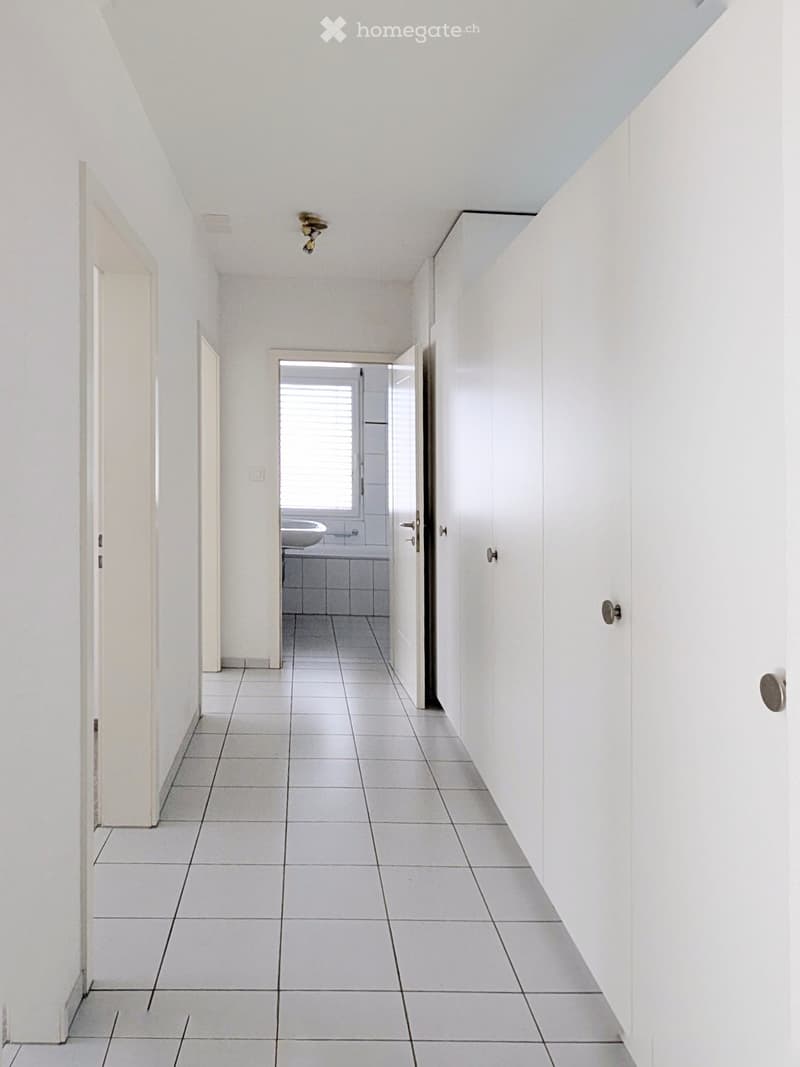 A clean, white-walled hallway with tiled flooring, leading to a bathroom visible at the end. The hallway features a recessed ceiling light and several doors along the walls.