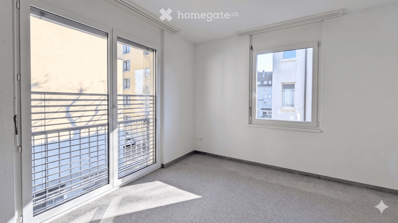 Empty room with white walls, grey carpeted floor, large window with white frame, and sliding glass door leading to a balcony with metal railing.