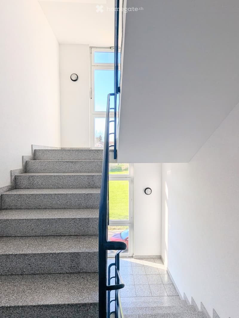 Modern staircase with grey granite steps, white walls, and a blue metal railing leading to a bright window area.