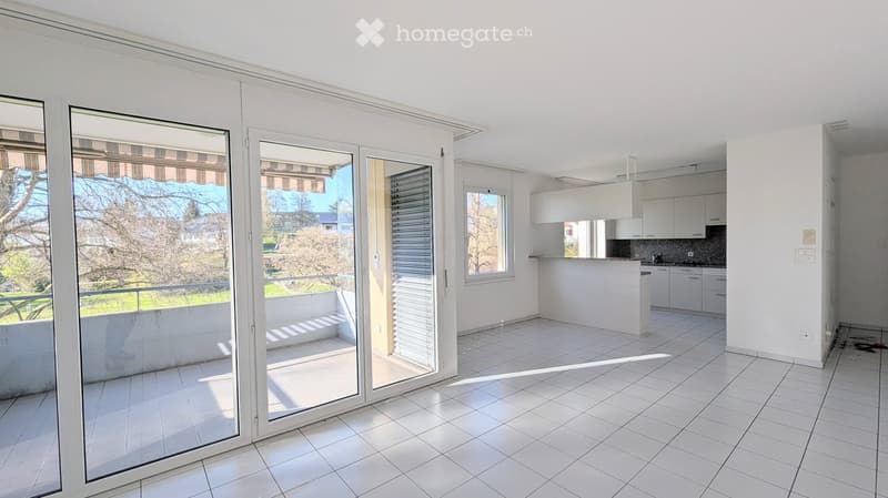 Bright, open-plan living and dining area with large sliding glass doors leading to a balcony, white tiled flooring, and a modern kitchen area with white cabinets and a countertop.