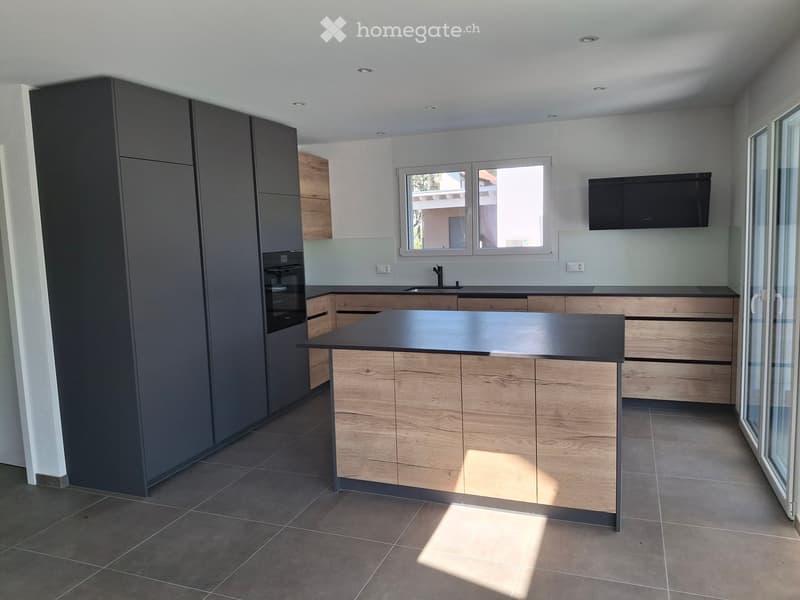 Modern kitchen with grey and light wood cabinets, central island, sink, and built-in oven. Large windows provide natural light, and a flat-screen TV is mounted on the wall.