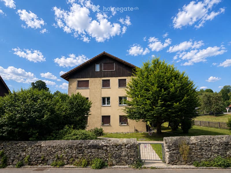 A two-story house with a pitched roof, beige walls, and wooden trim around the windows. The house is surrounded by greenery, including trees and shrubs, and is enclosed by a stone wall with a metal gate.