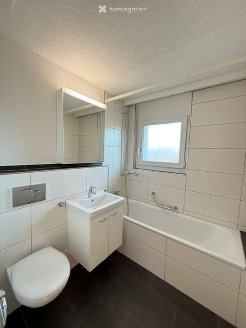 Modern bathroom with white tiles, a sink, toilet, bathtub, and a mirror above the sink. The room features a window with blinds and a sleek, contemporary design.