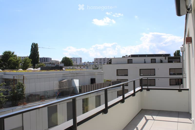 Balcony with metal railing, tiled floor, overlooking a modern building complex with rooftop greenery and surrounding urban structures.