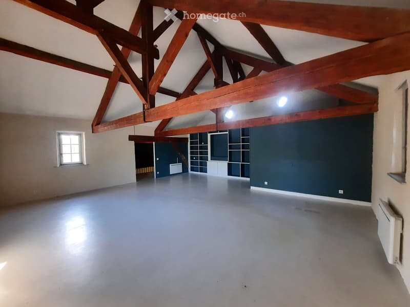 Empty room with exposed wooden beams, white walls, and a single window. The floor is smooth and grey, and there is a small built-in shelving unit in the corner.