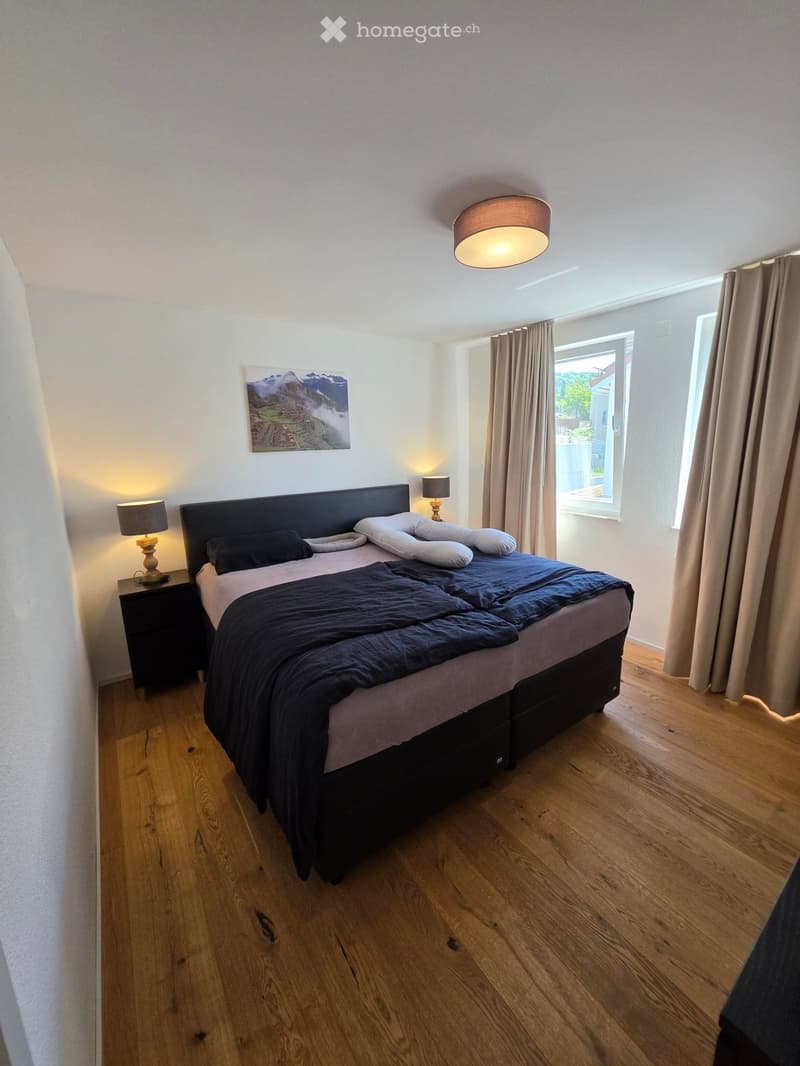 Modern bedroom with wooden flooring, large bed with dark bedding, nightstand with lamp, and a picture on the wall.