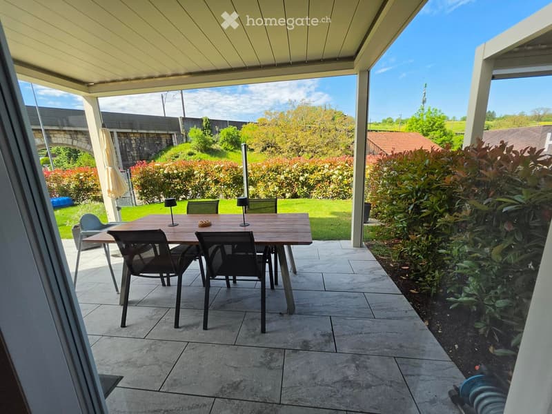 Patio area with a wooden table, chairs, and outdoor lighting, surrounded by greenery and a clear sky.