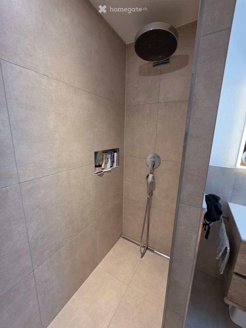 Modern bathroom with beige tiles, a handheld showerhead, a control valve, a small shelf with toiletries, and a towel hanging on a rack.