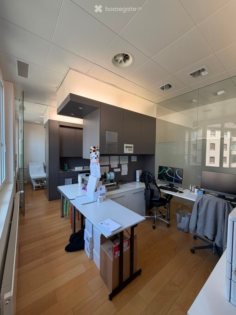 Modern office space with wooden flooring, white tables, chairs, laptop, computer screen, keyboard, cabinets, and a small sink area.