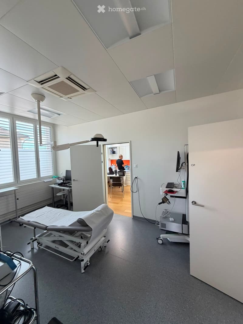 A modern medical examination room with an exam table, chairs, a screen, and a lamp. The room has large windows and a door leading to an adjacent office area.