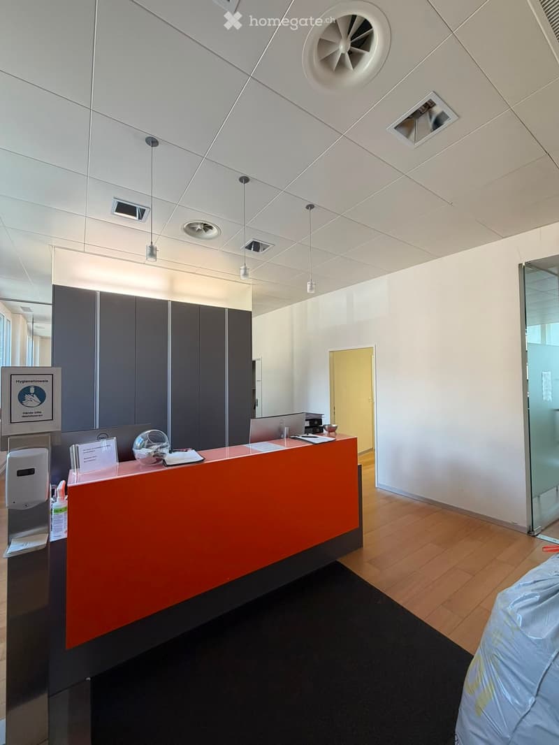 Modern reception area with a bright orange desk, laptop, hand sanitizer, and a sign on the wall. The ceiling features geometric patterns and recessed lighting.