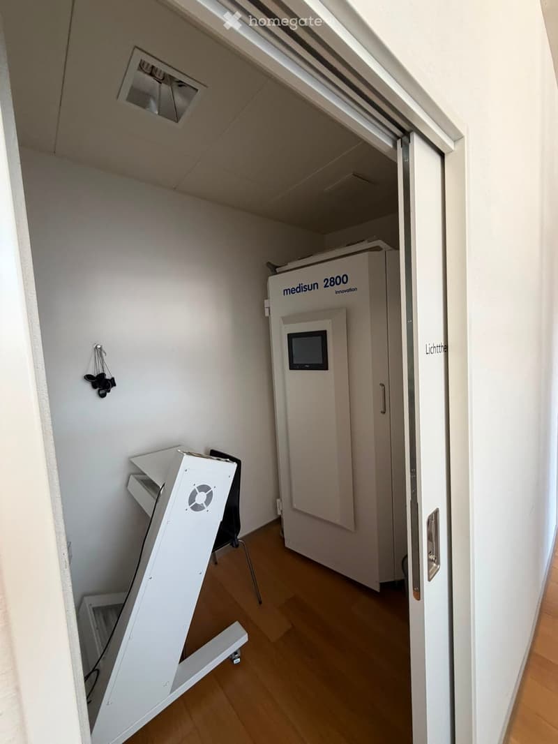 A small, well-lit room with a wooden floor, a white cabinet labeled "medisun 2800 Innovation" with a screen, and a white chair with a screen on a stand.
