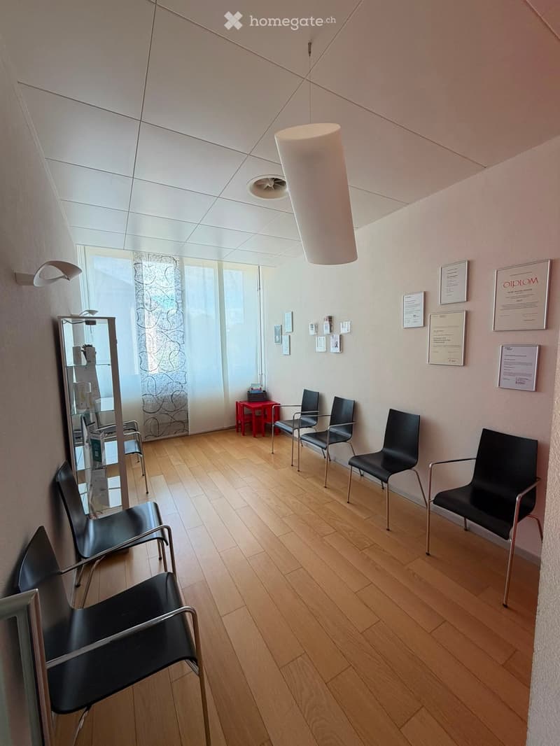 Waiting area with wooden floor, several black chairs, a small red table, a mirror, and framed certificates on the wall.
