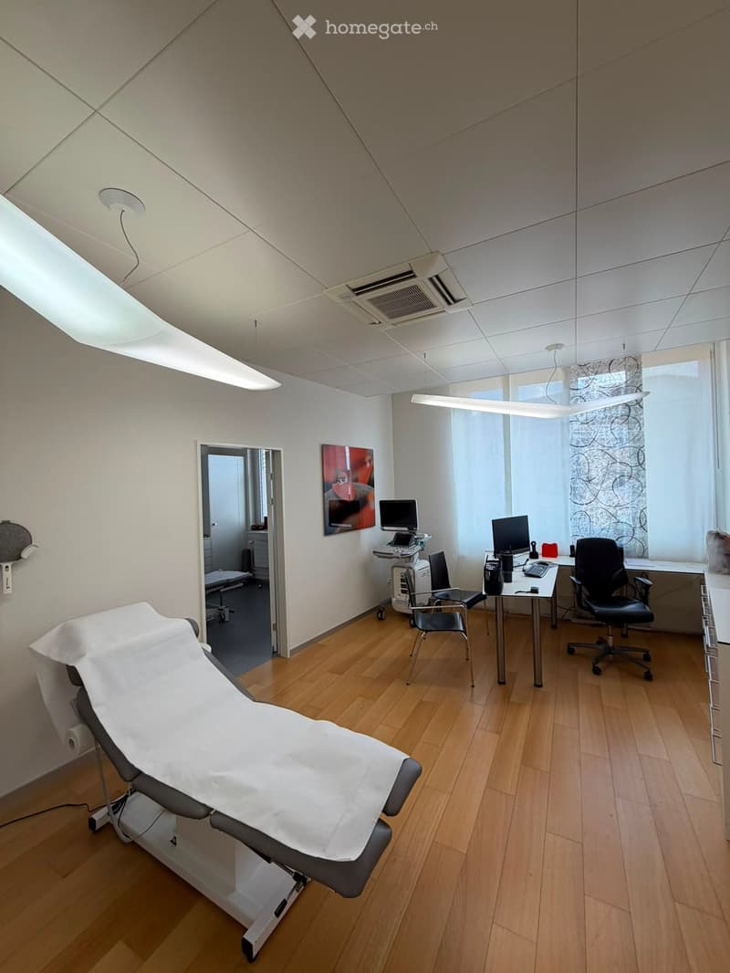 Modern medical examination room with wooden flooring, large windows, exam table, office furniture, and medical equipment.