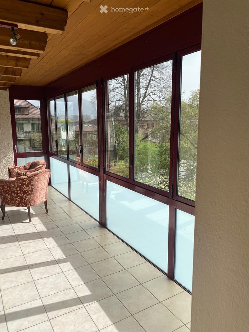 Bright, tiled balcony with large glass windows, wooden ceiling, and a single floral-patterned chair.
