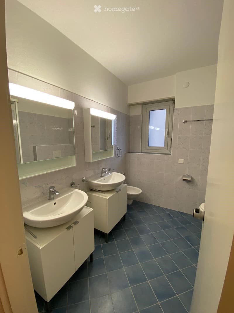 Modern bathroom with two sinks, mirrors, cabinets, and a toilet. Blue tiled floor and white walls.