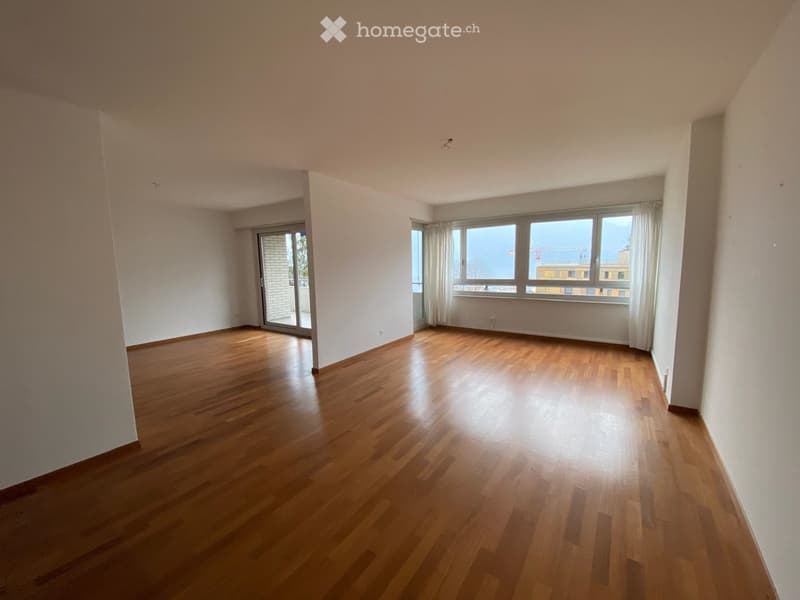Empty room with wooden flooring, white walls, large windows with curtains, and a sliding glass door leading to a balcony.