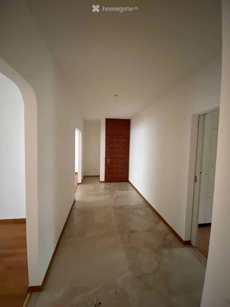 Empty hallway with white walls, wooden baseboards, and a tiled floor. Features a closed wooden door and a partially visible adjacent room.