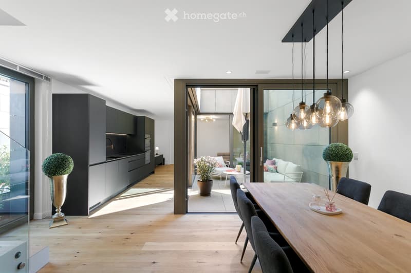 Modern open-plan dining and living area with light wood flooring, large glass doors leading to a patio, pendant lights, a dining table with chairs, a sofa, and decorative vases.